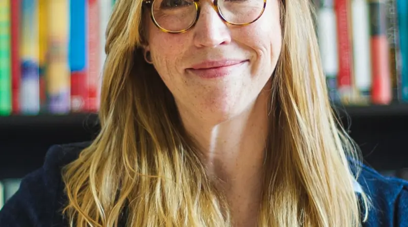 A woman with blonde hair and glasses smiles at the camera in front of a bookshelf.