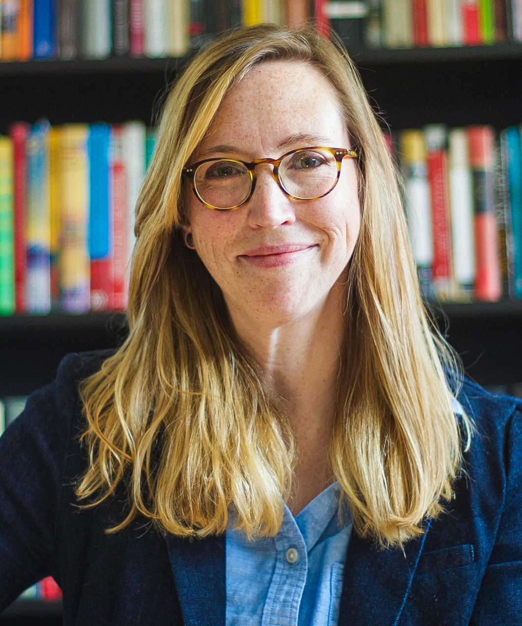 A woman with blonde hair and glasses smiles at the camera in front of a bookshelf.